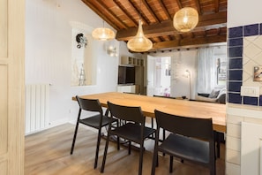 A dining area with a wooden table and four chairs, a hanging light fixture, and a view into another room with a sofa and a television.