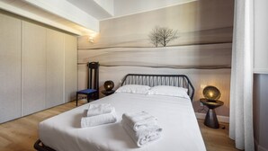 A neatly made bed with folded towels, a bedside table with a lamp, and a chair in a modern room with a wooden floor and a wall with a tree silhouette. - Giglio Celeste apartment in Florence by Mmega (Firenze)