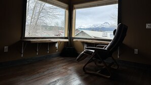 Une pièce dotée d’un plancher en bois, d’un fauteuil à bascule et de deux grandes fenêtres donnant sur un paysage de montagne et de forêts.