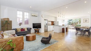 A spacious living area with a sofa, coffee table, and a black chair. A kitchen with a dining area is visible in the background. - Lilly`s Cottage - 3bdrm Heritage House, South Fremantle (South Fremantle)
