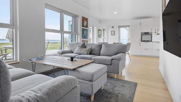 A modern living room with a grey sofa set, a wooden coffee table, and a view of the outdoors through large windows. - Stunning home in Holbæk with kitchen (Holbæk)