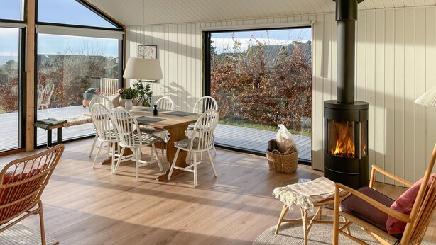 A cozy room with a wooden table, white chairs, a wicker chair, a sofa, and a black wood-burning stove.