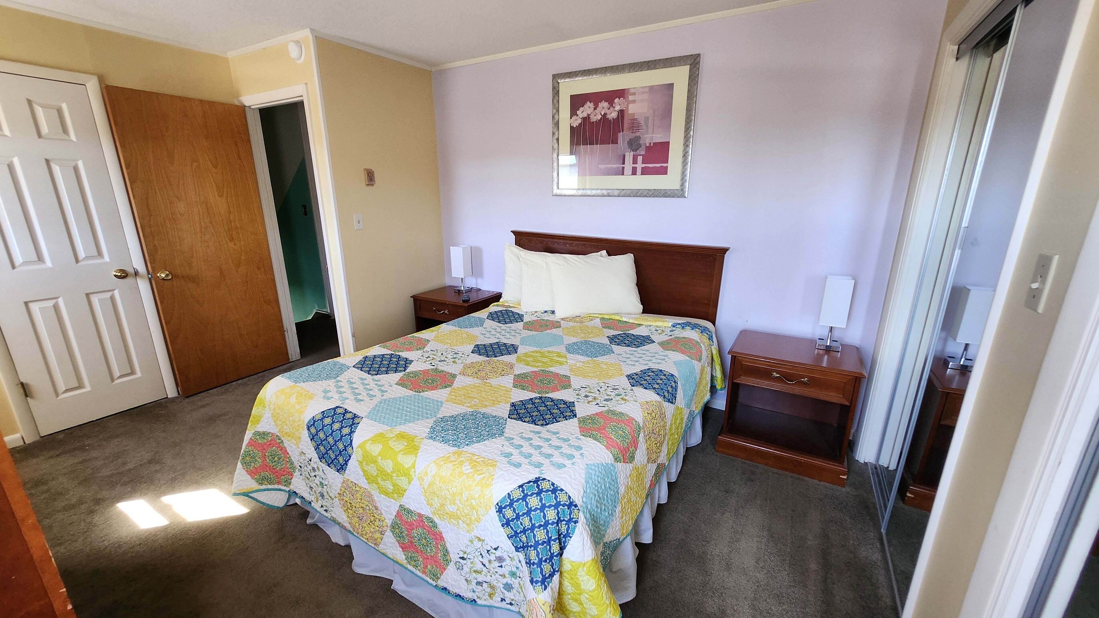 Ein Hotelzimmer mit einem Bett, einem Nachttisch, einem Tischchen neben dem Bett, einem gerahmten Bild an der Wand und einer Tür.