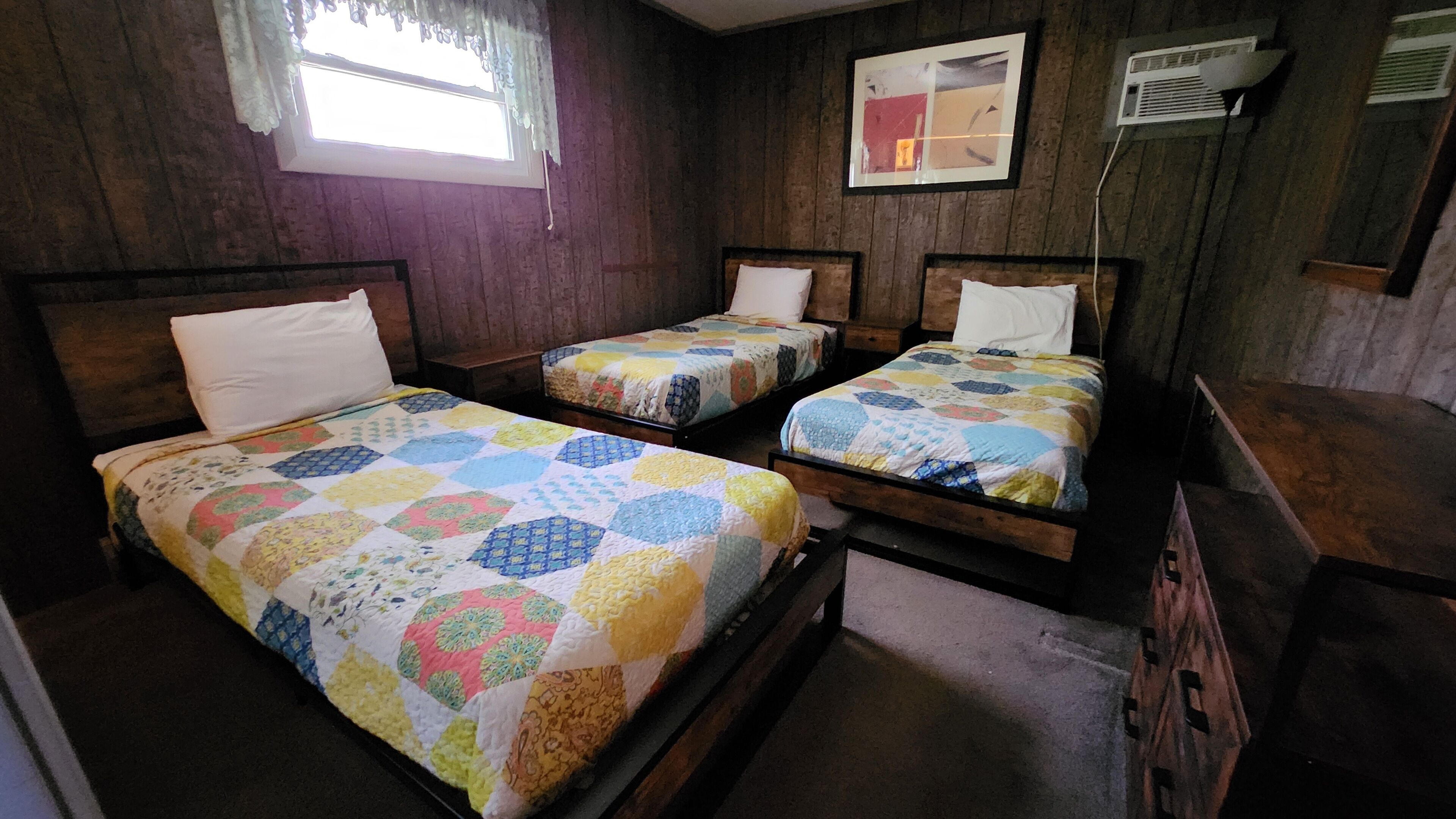 Una habitación con tres camas de madera, cada una con un colcha de colores. Se observa una ventana con cortinas de encaje y un cuadro en la pared.