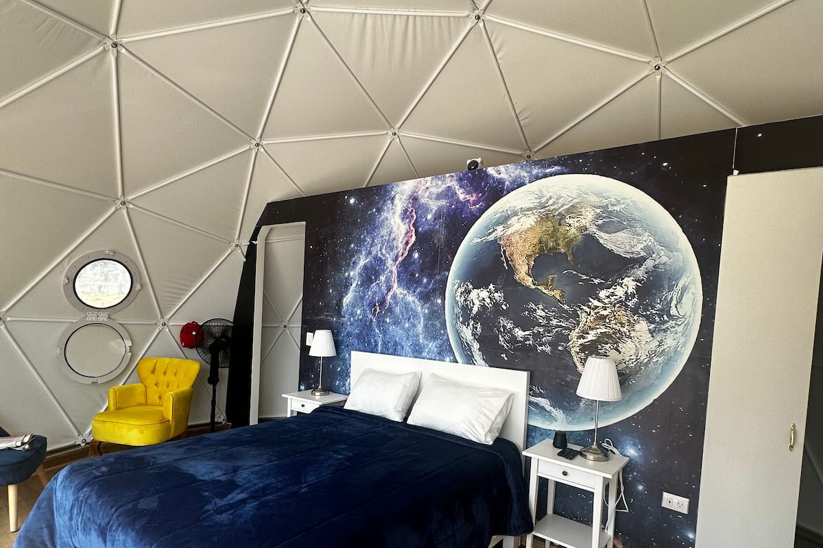 A bedroom with a geometric ceiling design, a bed with a blue bedspread, a yellow armchair, a round mirror, and a wall mural of Earth.