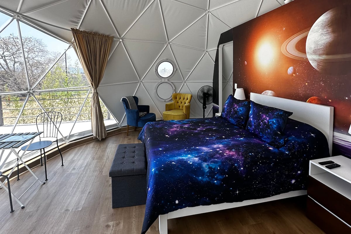 A bedroom with a galaxy-themed bedspread, a round window, and a view of the outdoors.