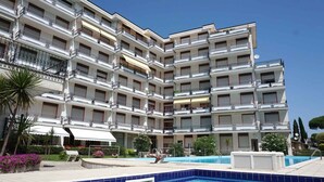 A multi-story hotel with balconies, a swimming pool, and outdoor seating areas. - Holiday apartment with oceanfront pool (Porto Santa Margherita)
