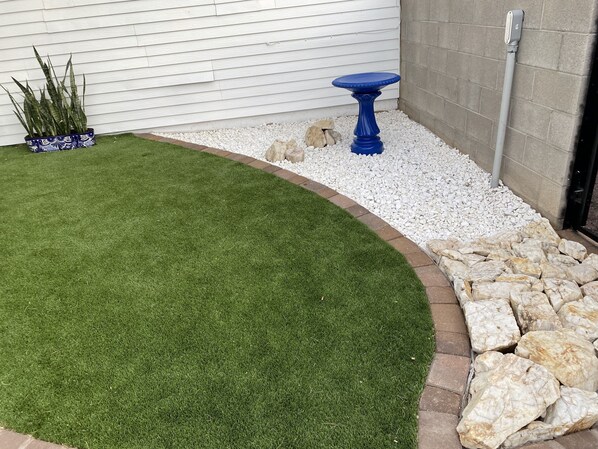 Área de jardim ajardinada com grama sintética, uma mesa azul e pedras decorativas.