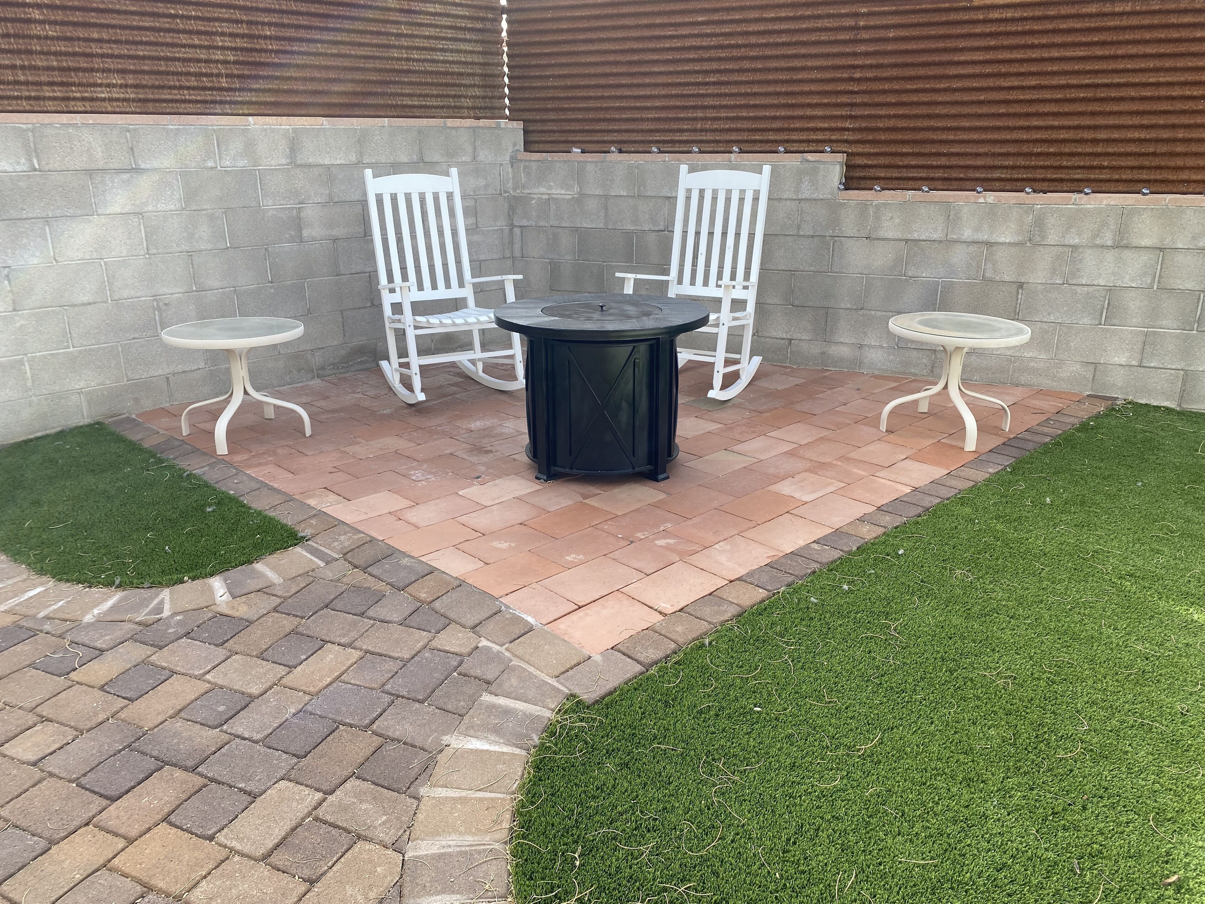 Eine Terrassenfläche mit Backsteinboden, zwei weiße Schaukelstühle, ein runder Tisch und künstlicher Rasen.