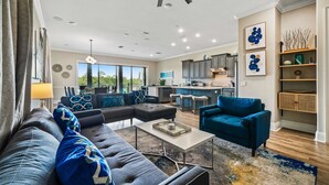 A modern living room with a sectional sofa, a blue armchair, and a coffee table. There is a kitchen area with bar stools and a dining table.