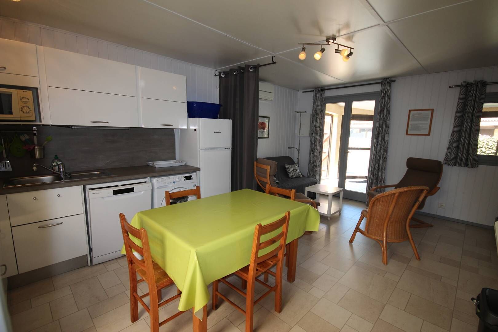 A compact kitchen with a dining area, a small table, and chairs. There is a refrigerator, a microwave, and a sink. The room has a window with curtains and a door leading outside.