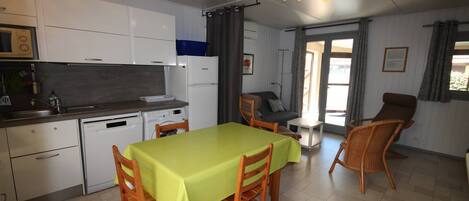 A compact kitchen with a dining area, a small table, and chairs. There is a refrigerator, a microwave, and a sink. The room has a window with curtains and a door leading outside.