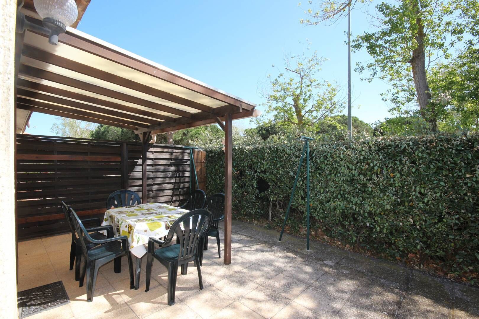 A covered patio area with a table and chairs, a pergola, and a hedge in the background.