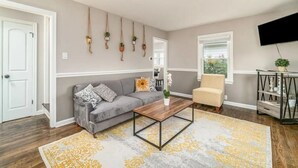 Ein Wohnzimmer mit grauem Sofa, Couchtisch, gemustertem Teppich, Sessel, Fernseher und einer wandmontierten Pflanzenanordnung.