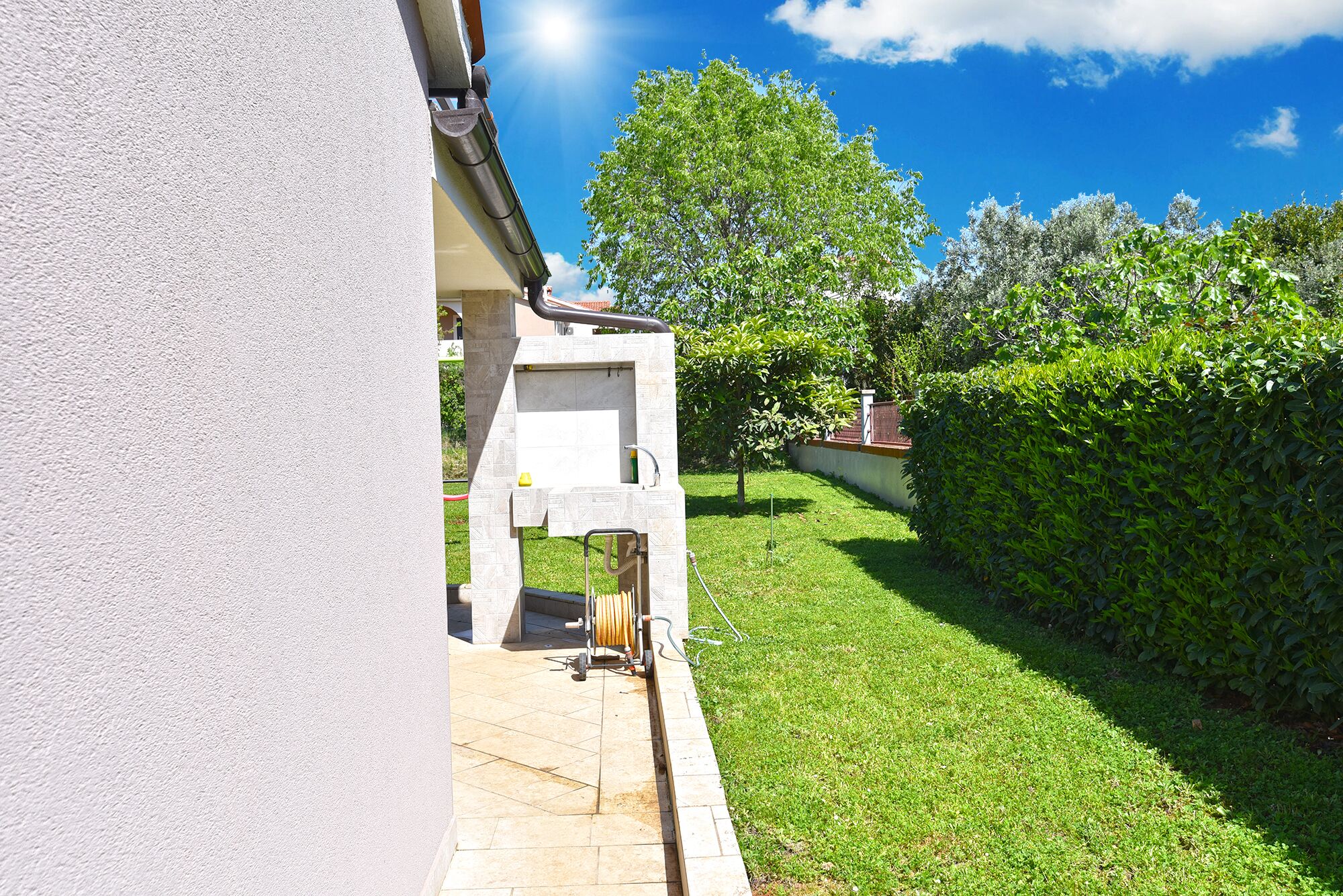 Un jardin bien entretenu, avec un chemin pavé menant à l’entrée d’un bâtiment, entouré d’une végétation luxuriante et surplombé d’un ciel bleu clair.