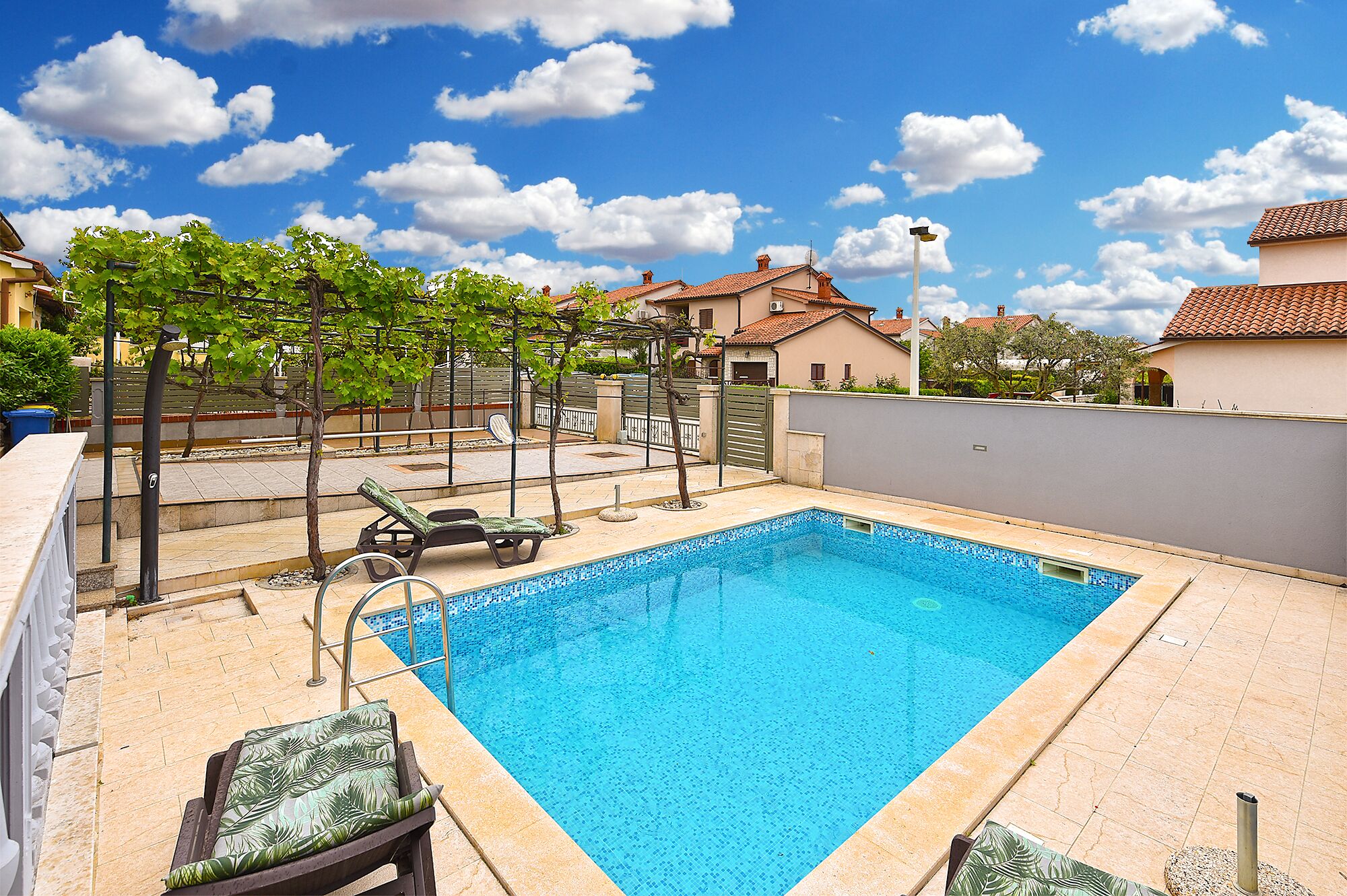 Une piscine rectangulaire avec une terrasse carrelée, entourée de chaises longues et d’une pergola couverte de plantes grimpantes.
