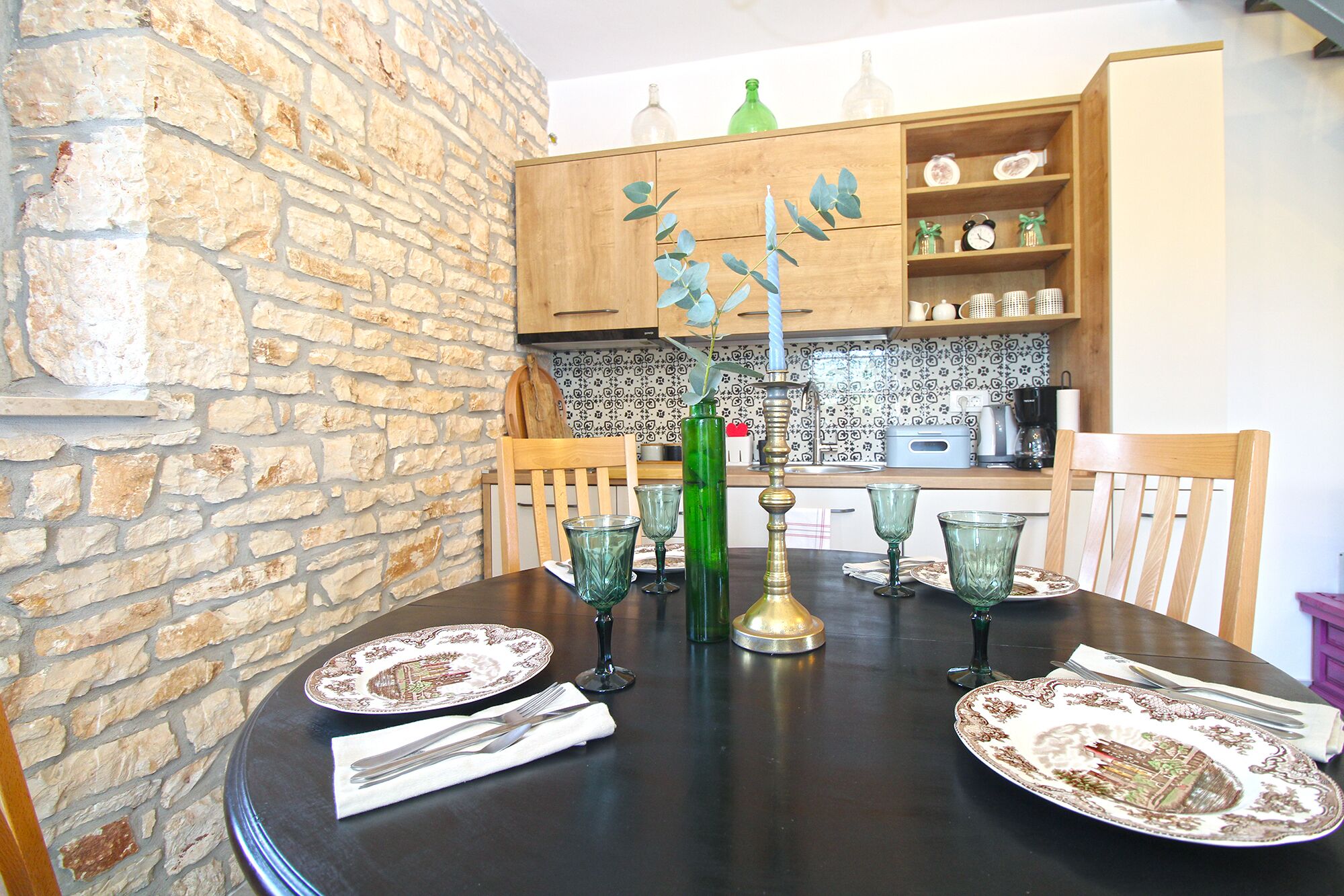 A dining table set with plates, glasses, and cutlery, with a brick wall and wooden cabinets in the background.