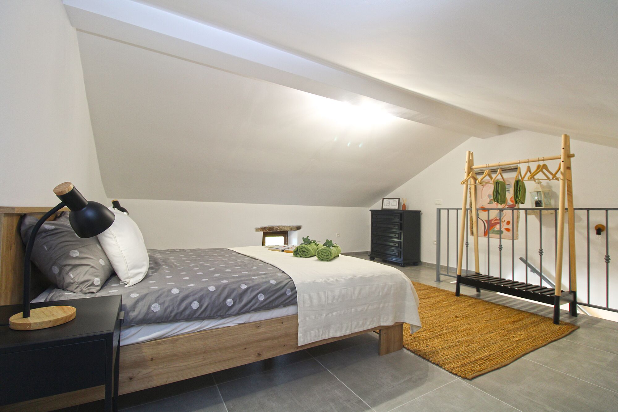 A bedroom with a sloped ceiling, a bed with a grey and white polka dot bedspread, a wooden nightstand, a black lamp, a small table with a lamp, a black dresser, and a hanging clothes rack.