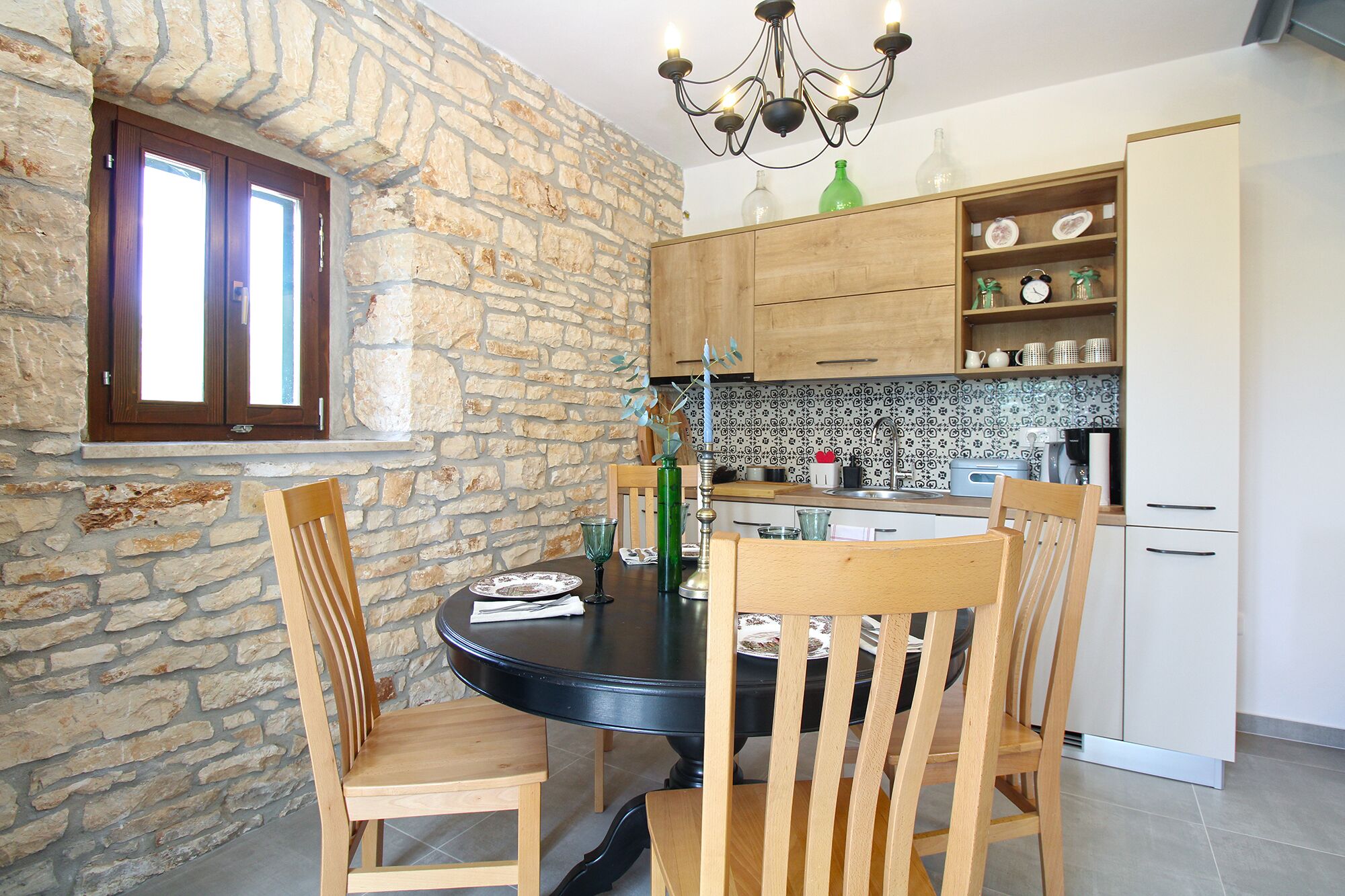 A dining area with a stone wall, wooden chairs, a round table, and a kitchen in the background.