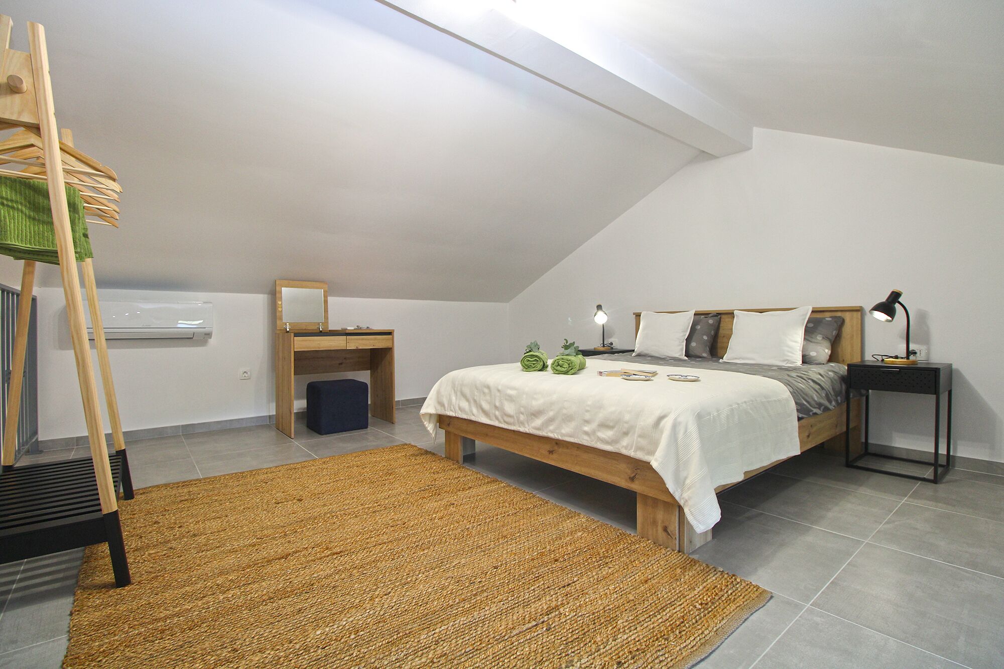 Une chambre à coucher comprenant un lit, un bureau avec un miroir, un petit tabouret, une table de chevet avec une lampe et un porte-vêtements suspendu.