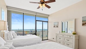 A hotel room with two beds, a ceiling fan, a large window with a city view, a white dresser, and a mirror.