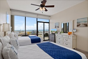 A hotel room with two beds, a ceiling fan, a large window with a view of the city, and a dresser with a mirror and a painting.