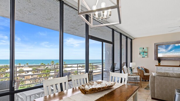 A spacious living area with a large window overlooking a beach, a wooden dining table, and a comfortable seating arrangement.