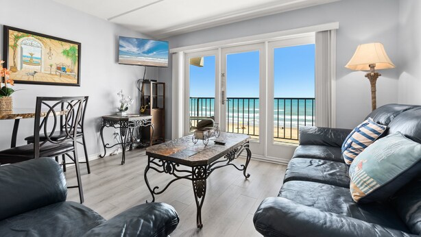 Sala de estar com vista para a praia, área de jantar com mesa e cadeiras, e sofá com almofadas decorativas.