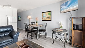 Uma sala de estar moderna com um sofá de couro preto, uma mesa de jantar com cadeiras, uma TV montada na parede e um quadro com uma cena de praia.