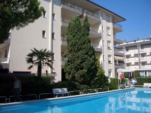 A swimming pool with a bench, a tall tree, and a multi-story building with balconies.