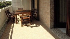 Een overdekt terras buiten met een houten tafel en stoelen, een gele parasol en uitzicht op de stad.