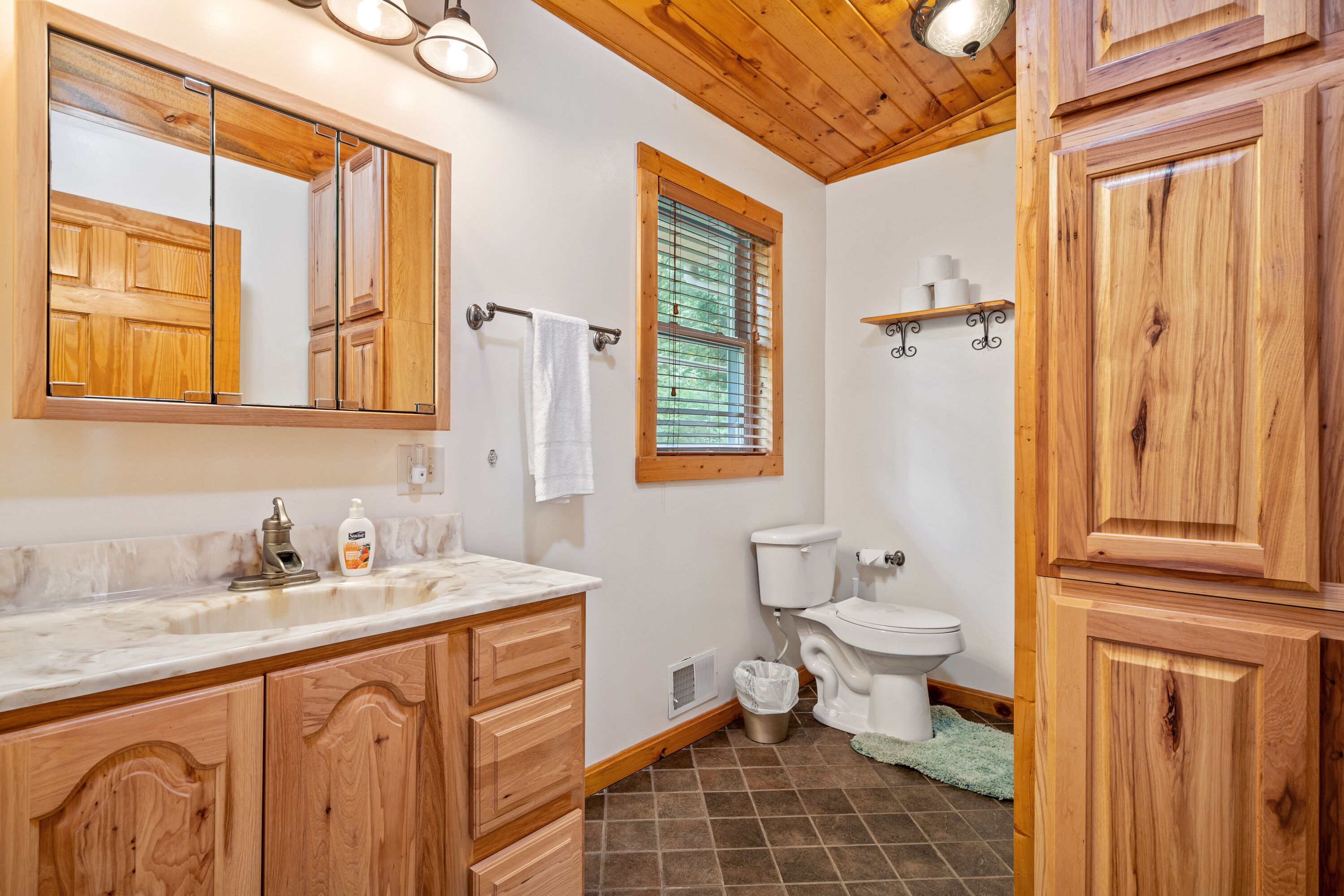A bathroom with wooden cabinets, a marble sink, a toilet, and a window with blinds.