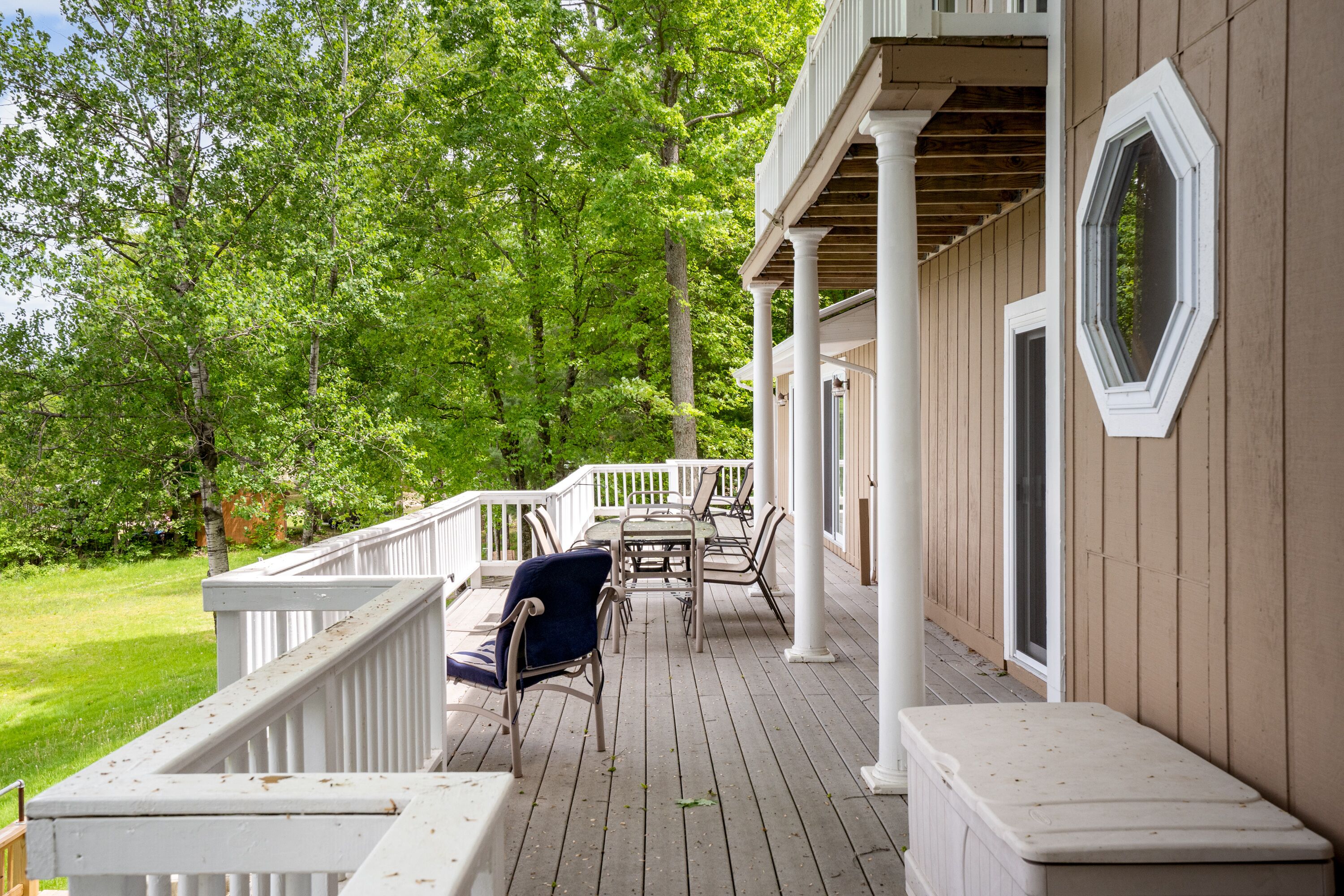 Una terraza de madera con barandillas blancas, mobiliario exterior y vista a un denso bosque verde.