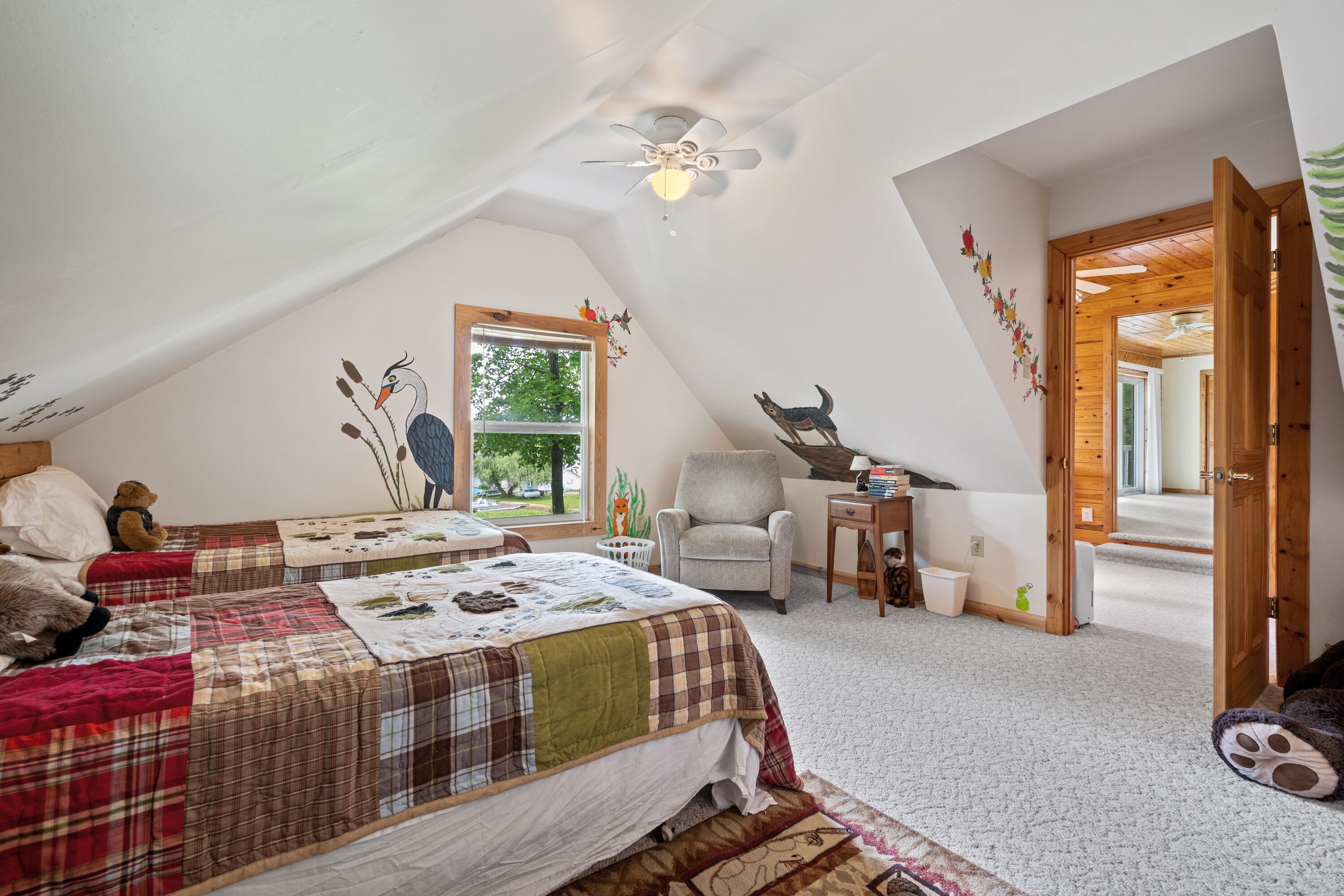 A bedroom with a sloped ceiling, a bed with a plaid bedspread, a chair, a desk, and a window with a view of trees.