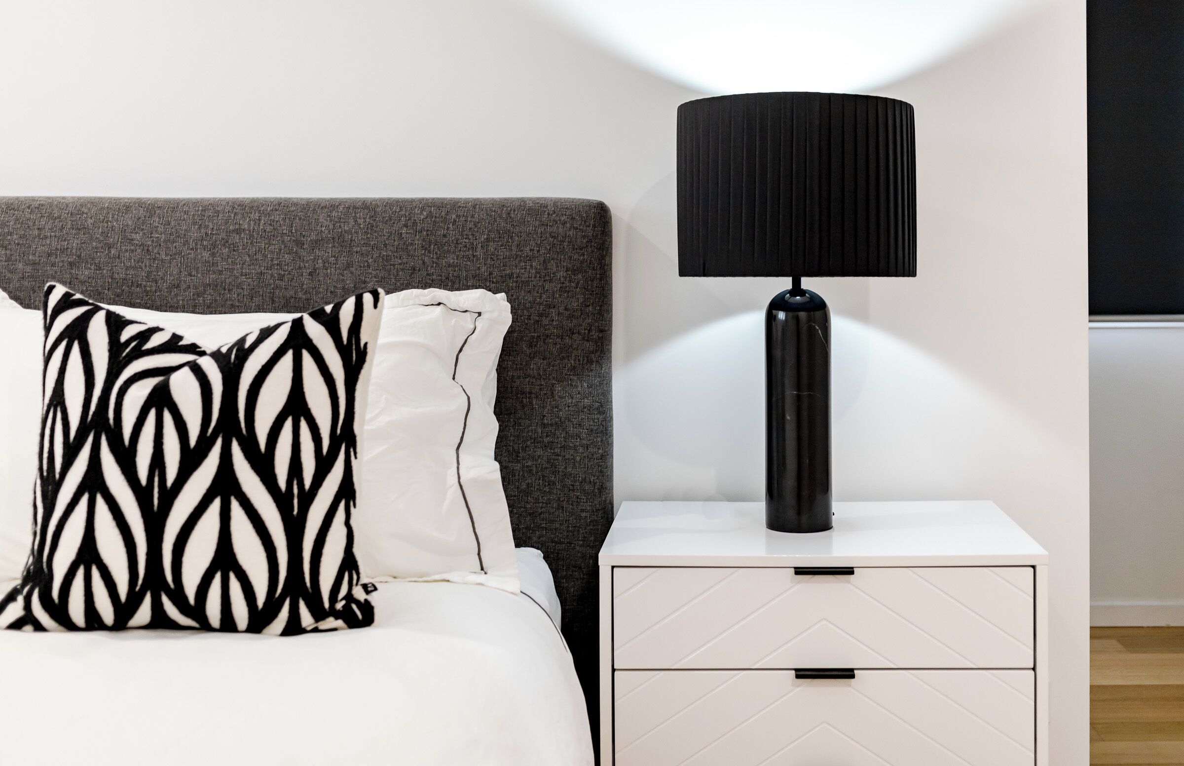 Een opgemaakt bed met gestreepte kussens, een bedlampje en een witte nachtkastje met lades.