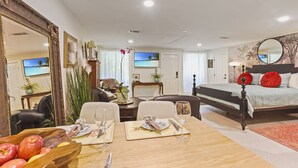 Una habitació d'hotel amb un llit, una taula de menjador amb un plat de fruita i gots de vi, un televisor i un mirall gran.