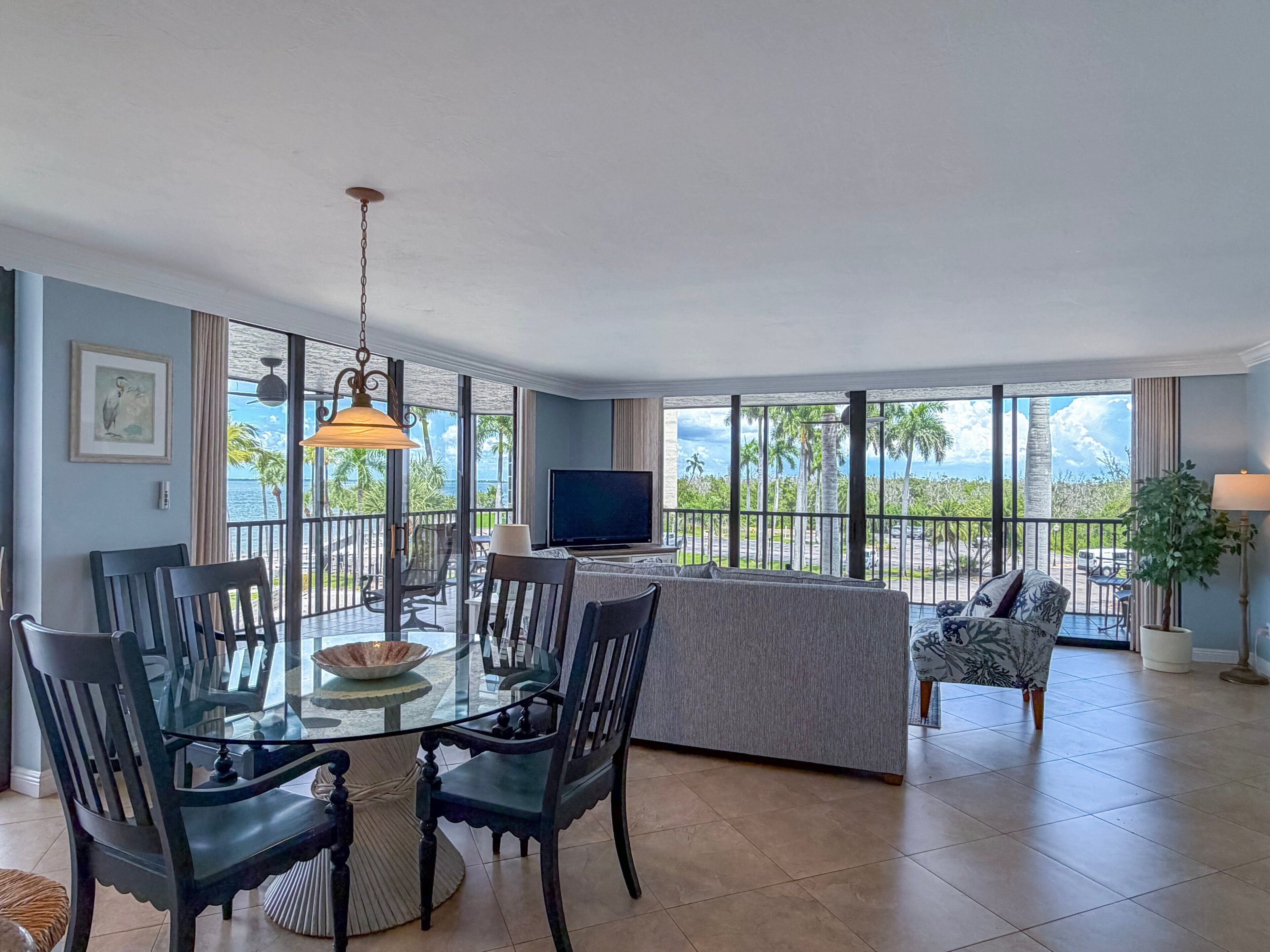 A hotel room with a balcony, a dining area, a TV, and a large window offering a view of palm trees and the ocean.