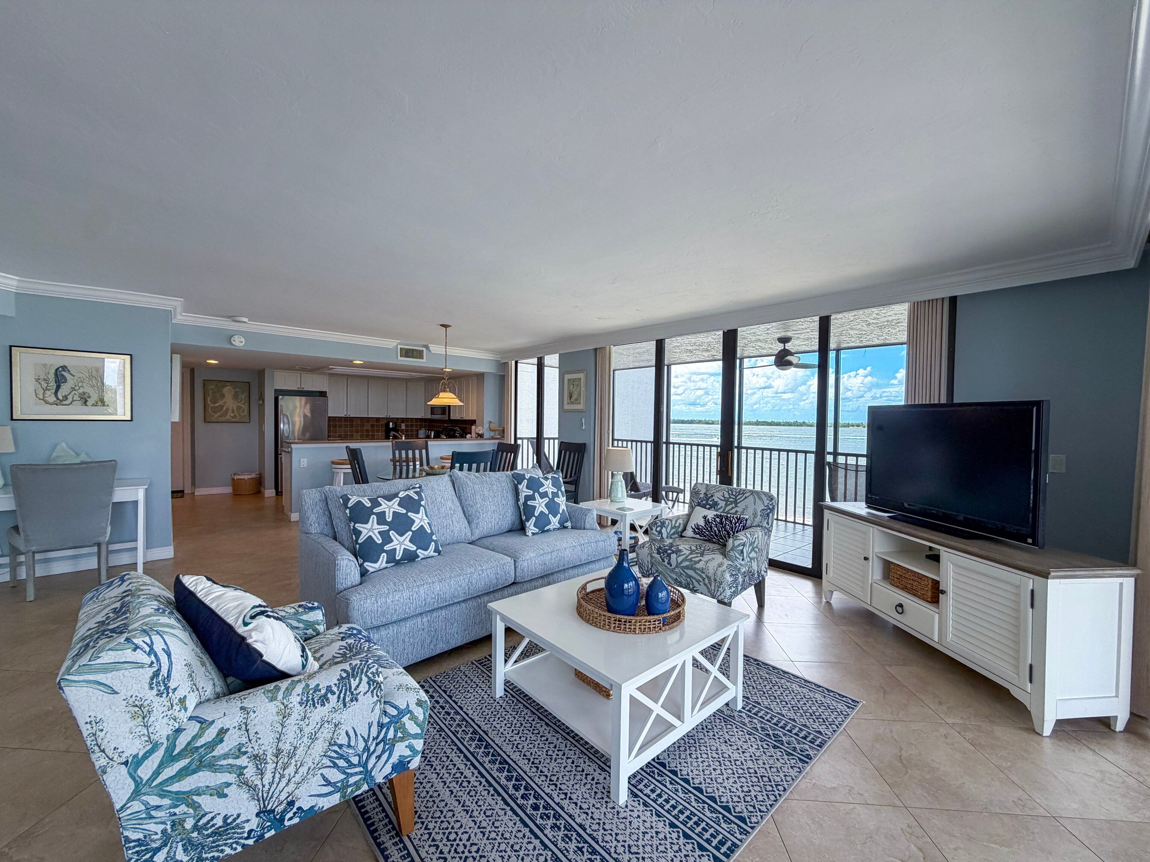 A living room with a sofa, armchairs, a coffee table, and a TV. There is a balcony with a view of the water.