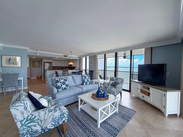 Una sala de estar con sofá, sillones, una mesa de centro y televisor. Hay un balcón con vista al agua.