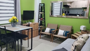 Un espace de vie compact comprenant un coin repas, un canapé, un téléviseur et une cuisine visible à travers une fenêtre.