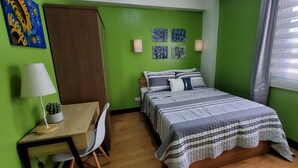 Une chambre à coucher avec un lit, un bureau, une chaise, une lampe, un cactus et des œuvres d’art mural.
