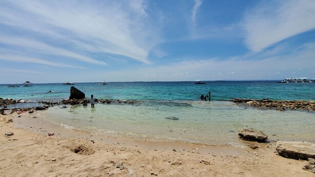 Une plage avec une eau claire, des rochers et des bateaux visibles au loin.