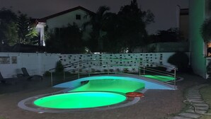 Une piscine dont l’eau est illuminée de lumière verte, entourée d’un chemin en briques et d’un mur blanc agrémenté de plantes en pot.