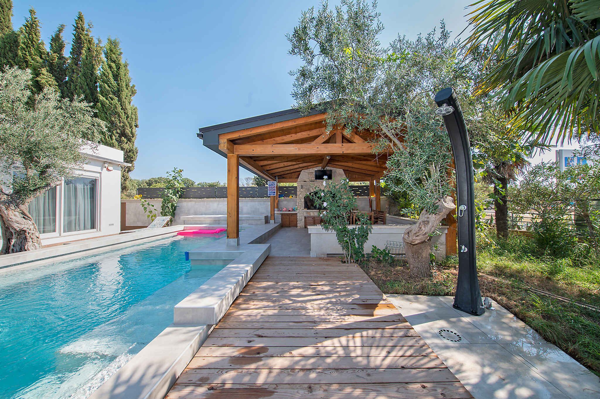 A wooden deck leading to a covered outdoor area with a pool, surrounded by trees and greenery.
