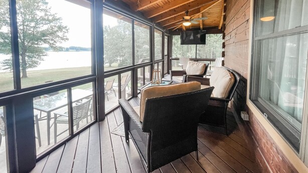 A covered outdoor area with wooden flooring, wicker furniture, and a large window offering a view of a lake. - Waverly Lakehouse - private cabin, lake view, close to 
Old Waverly/Mossy Oak (West Point)