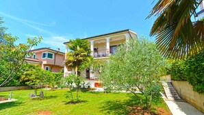 A two-story building with a balcony, surrounded by a well-maintained garden with various plants and a palm tree. - Holiday apartment with air conditioning and barbecue facilities (Poreč)