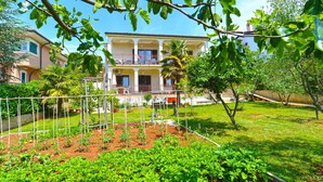 A two-story building with a balcony, surrounded by a garden with various plants and a fenced vegetable garden. - Holiday apartment with air conditioning and barbecue facilities (Poreč)