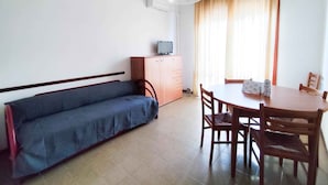 A room with a sofa, a small table with chairs, a TV on a cabinet, and a window with curtains. - Holiday apartment (Lido di Jesolo)