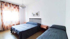 A bedroom with two beds, a nightstand, a hanging light, and a window with curtains. - Holiday apartment (Lido di Jesolo)