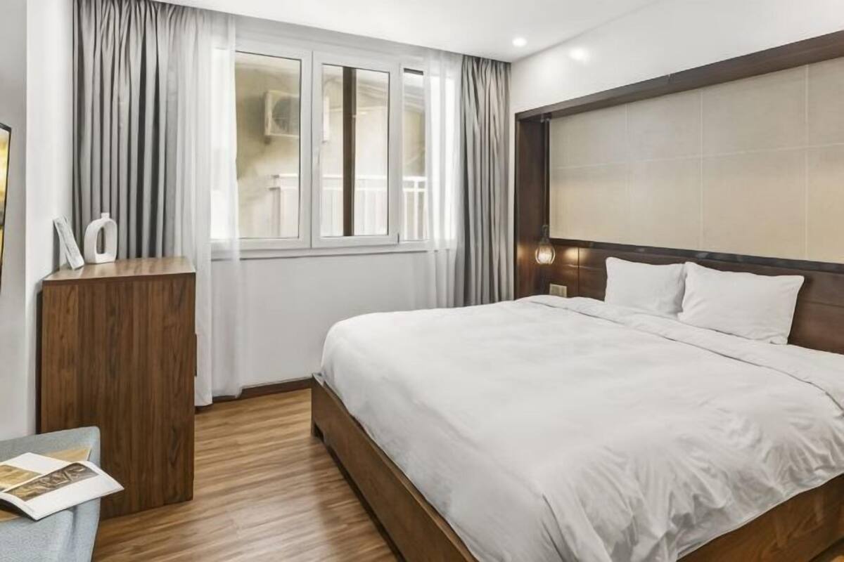 Une chambre d’hôtel avec un grand lit, une tête de lit en bois, un bureau avec une lampe et une fenêtre avec des rideaux.